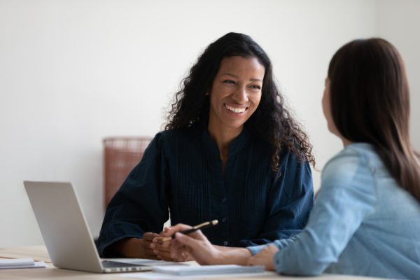 Smiling African American businesswoman talking to colleague, diverse employees networking, sitting at table in office, manager consulting client, using laptop, mentor coach training intern,
