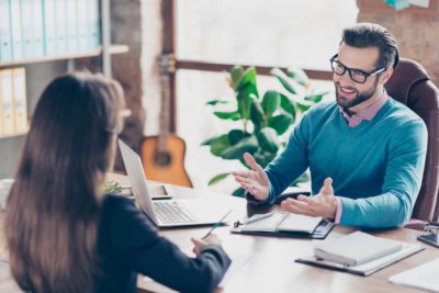 Job interview - Joyful, successful businessman asking candidate questions, sitting at desk in workplace on chair, girl making notes