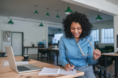 Smiling young businesswoman sitting alone at her desk in a large modern office writing ideas down in a notebook