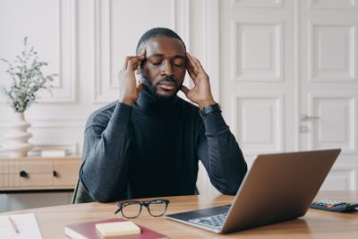 Frustrated tired afro american man office worker with closed eyes trying to concentrate while sitting at workplace, african ethnicity male employee massaging temples, suffering from headache at work