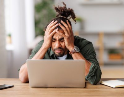 Frustrated tired young african american entrepreneur with closed eyes massages temples with fingers with painful expression on her face, sitting in front of laptop  working remotely online. Frustrated black guy has computer problem