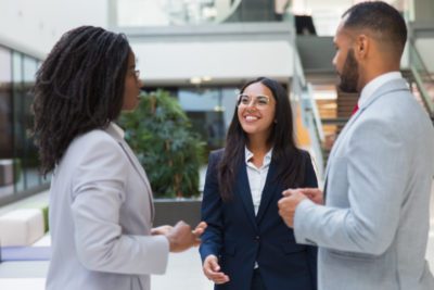 Friendly confident female business leader welcoming partners in office hall. Young business woman smiling and walking to diverse business couple. Cooperation concept