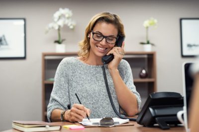 Beautiful mature woman talking on phone at creative office. Happy smiling businesswoman answering telephone at office desk. Casual business woman sitting at her desk making telephone call and taking notes on notebook.