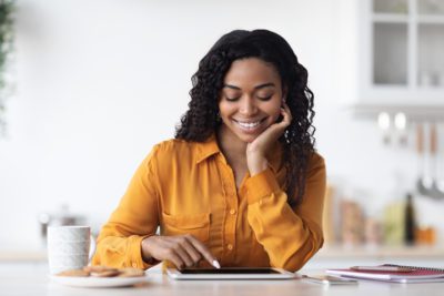 Happy millennial african american lady with curly hair SMM manager working from home, sitting at kitchen table, drinking coffee with cookies, using digital tablet and smiling, copy space